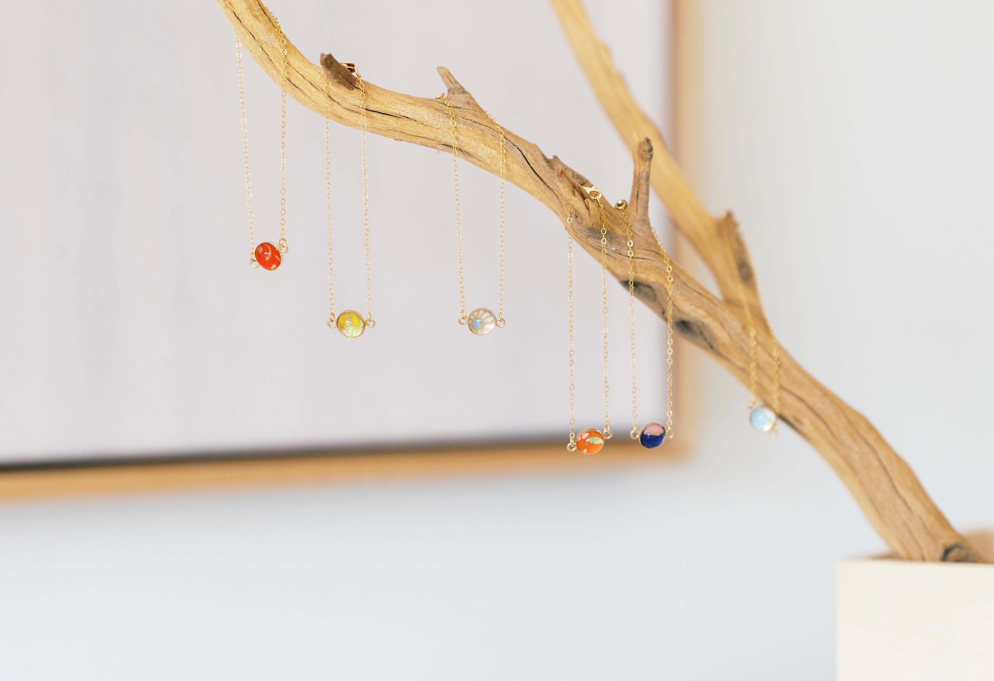A set of delicate gold-tone bezel bracelets, hanging from a decorative driftwood branch stand in a white pot against a bright, softly blurred background.