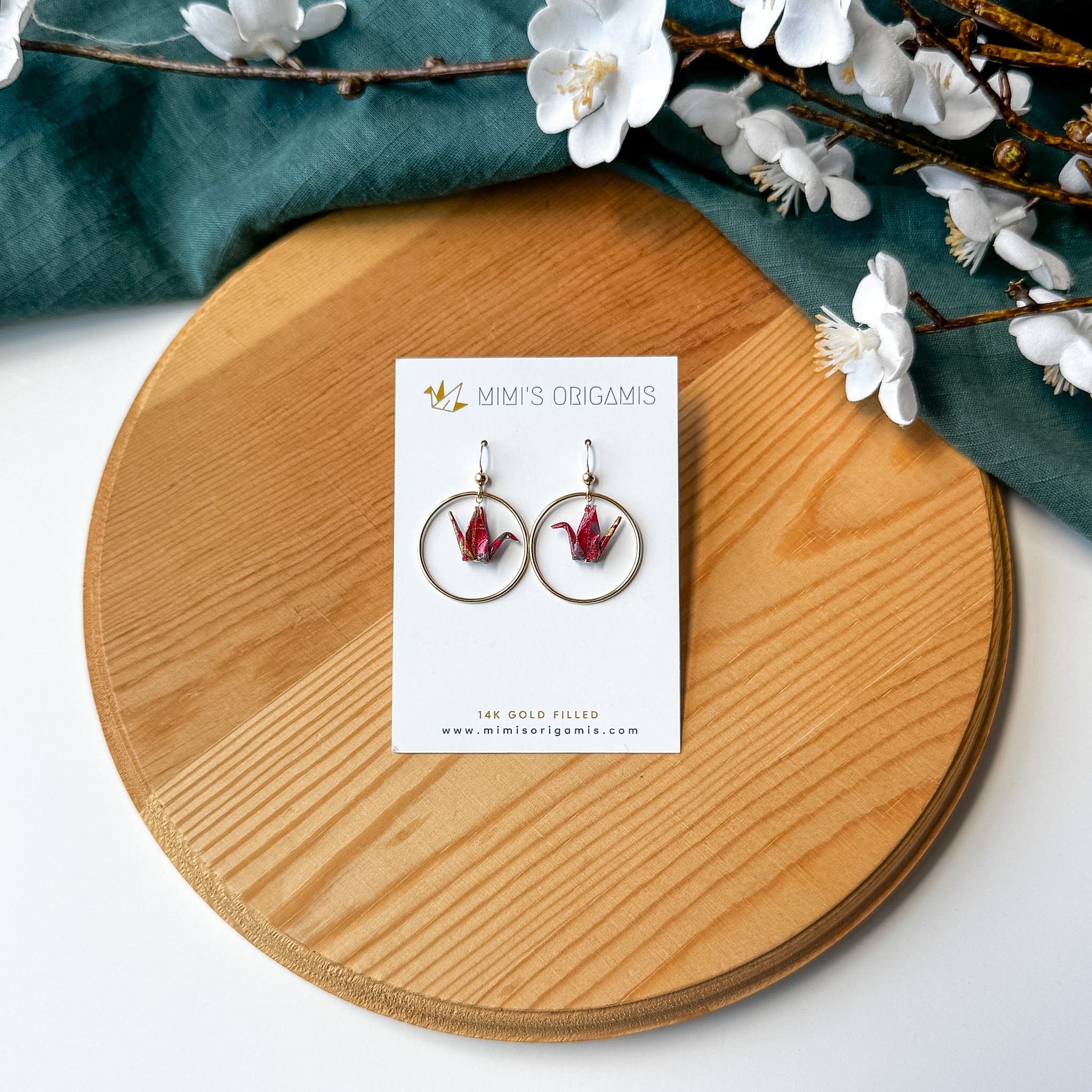 A pair of gold hoop earrings with red origami cranes inside, displayed on a white brand card, set on a round wooden board with a teal cloth and white blossoms.
