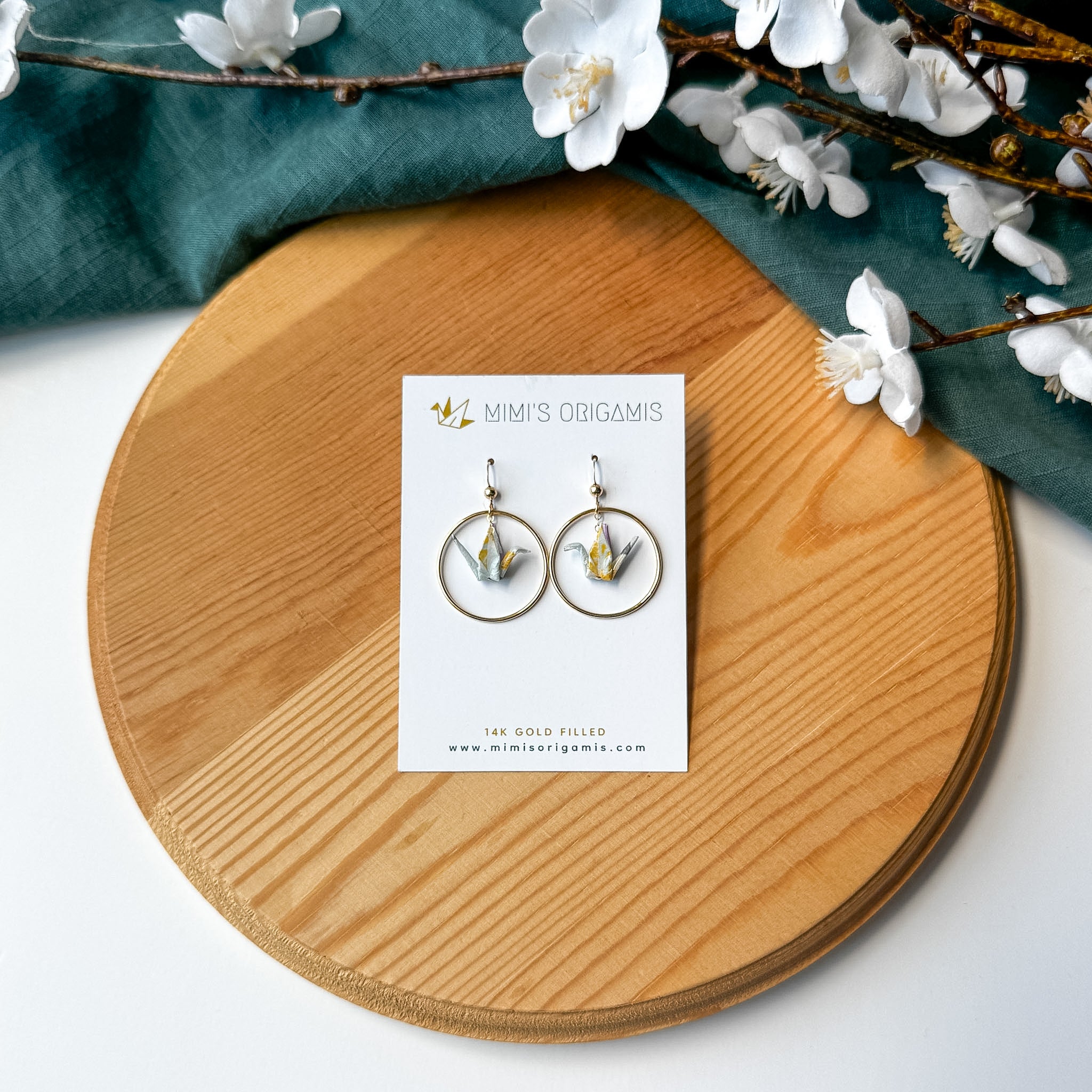A pair of gold hoop earrings with light blue and yellow origami cranes inside, displayed on a white brand card, set on a round wooden board with a teal cloth and white blossoms.