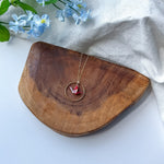 A gold-toned necklace with a circular pendant and a small red origami crane, displayed on a curved wooden board with light blue flowers and a white fabric backdrop.