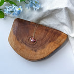A gold-toned necklace with a circular pendant and a small pink origami crane, displayed on a curved wooden board with light blue flowers and a white fabric backdrop.