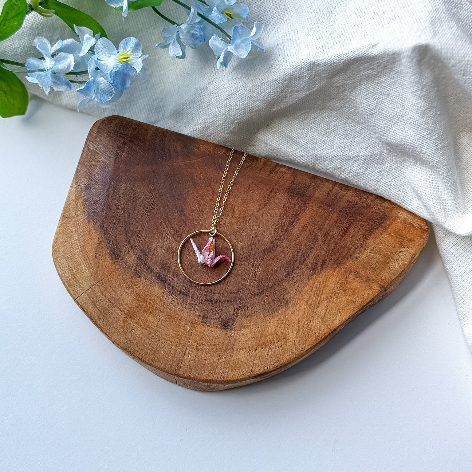 A gold-toned necklace with a circular pendant and a small pink origami crane, displayed on a curved wooden board with light blue flowers and a white fabric backdrop.