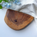 A gold-toned necklace with a circular pendant and a small indigo origami crane, displayed on a curved wooden board with light blue flowers and a white fabric backdrop.