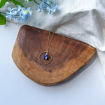 A gold-toned necklace with a circular pendant and a small blue origami crane, displayed on a curved wooden board with light blue flowers and a white fabric backdrop.