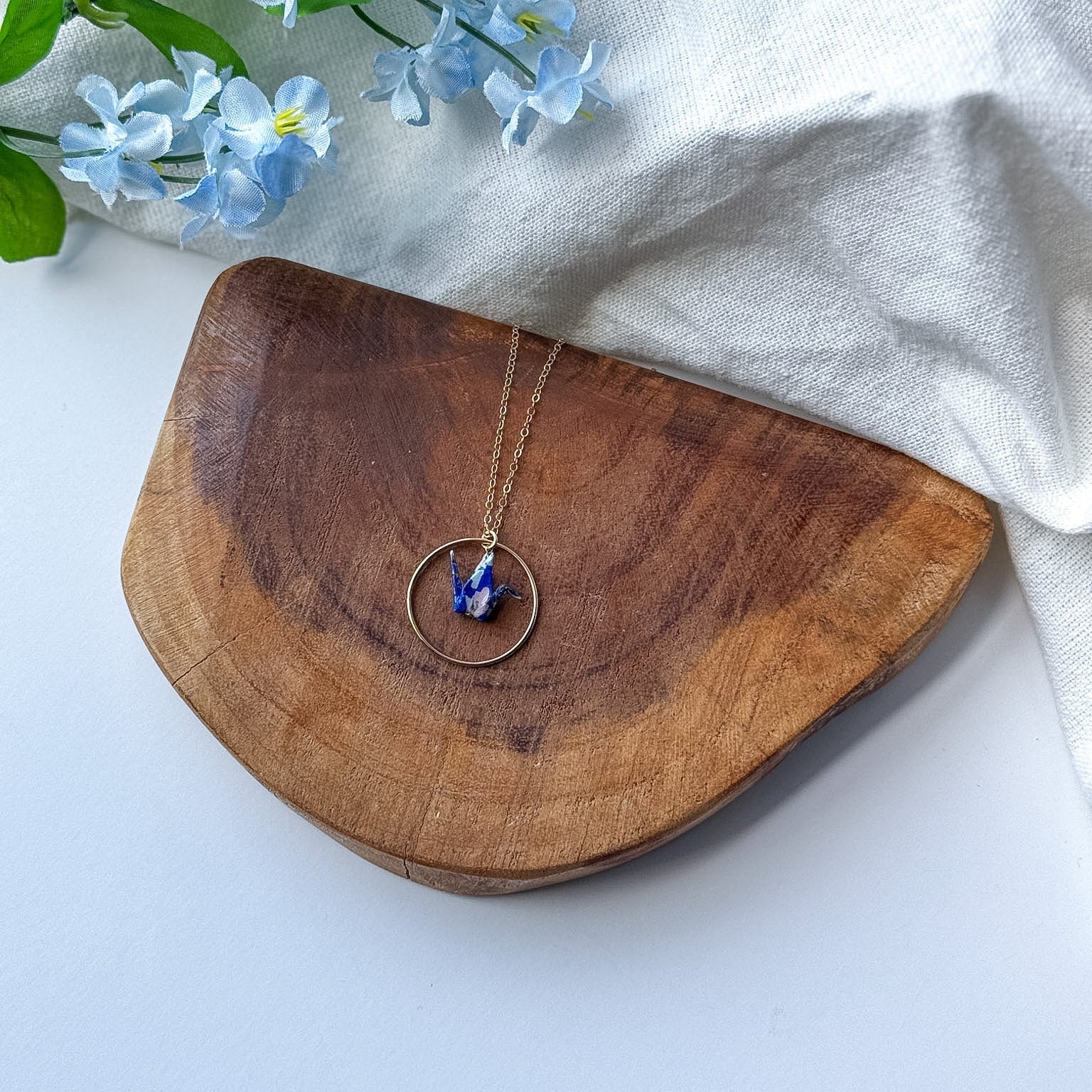 A gold-toned necklace with a circular pendant and a small blue origami crane, displayed on a curved wooden board with light blue flowers and a white fabric backdrop.