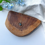 A gold-toned necklace with a circular pendant and a small teal origami crane, displayed on a curved wooden board with light blue flowers and a white fabric backdrop.
