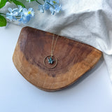 A gold-toned necklace with a circular pendant and a small teal origami crane, displayed on a curved wooden board with light blue flowers and a white fabric backdrop.