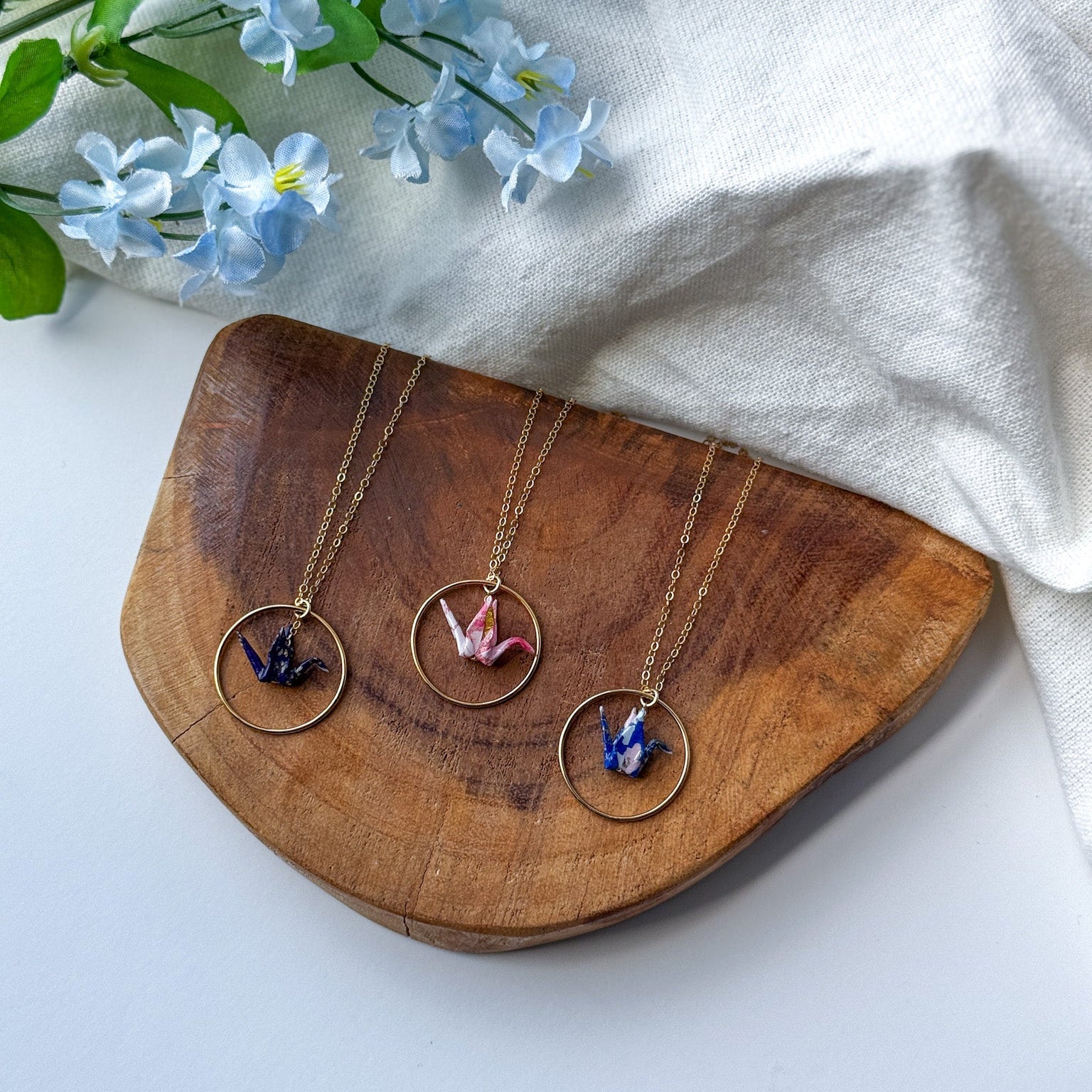 Three gold-tone necklaces with circular pendant frames and colorful crane pendants are arranged on a wooden tray, with light blue flowers and a white fabric backdrop.