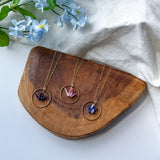 Three gold-tone necklaces with circular pendant frames and colorful crane pendants are arranged on a wooden tray, with light blue flowers and a white fabric backdrop.