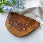 Three gold-tone necklaces with circular pendant frames and colorful crane pendants are arranged on a wooden tray, with light blue flowers and a white fabric backdrop.