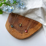 Three gold-tone necklaces with circular pendant frames and colorful crane pendants are arranged on a wooden tray, with light blue flowers and a white fabric backdrop.