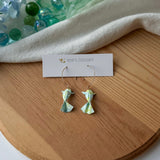 Pair of green origami koi fish earrings on a wooden surface with a white branded card