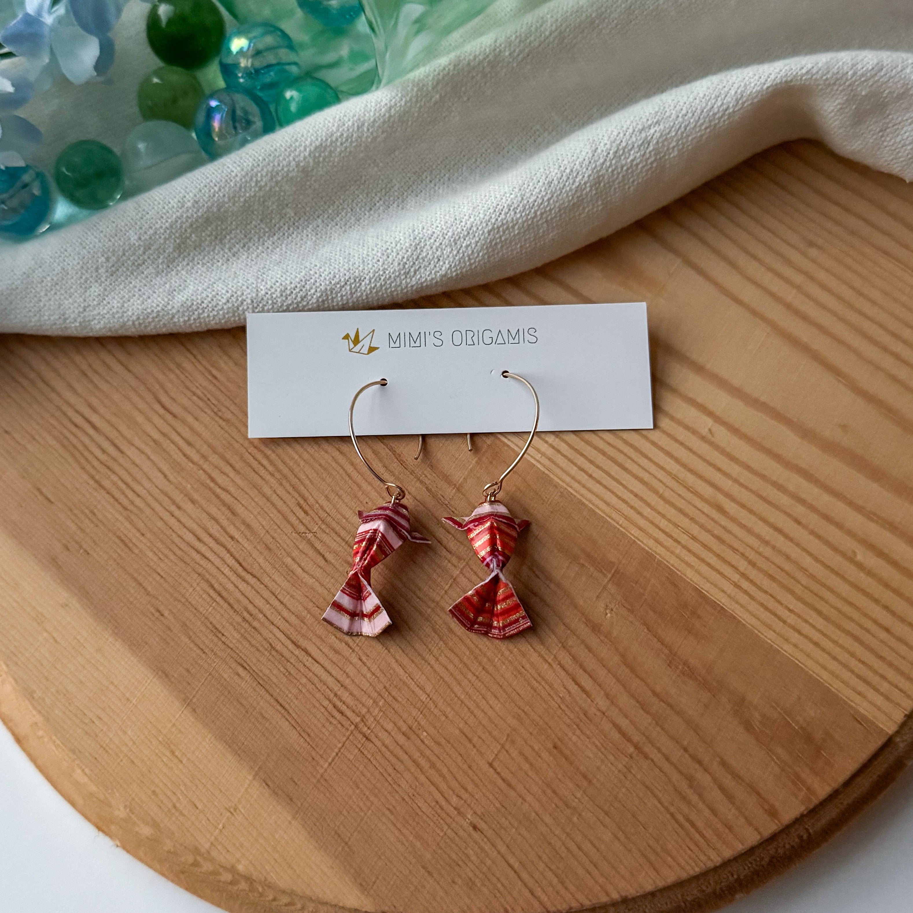 Pair of striped origami koi fish earrings on a wooden surface with a white branded card