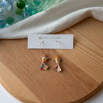 Pair of colorful origami koi fish earrings on a wooden surface with a white branded card