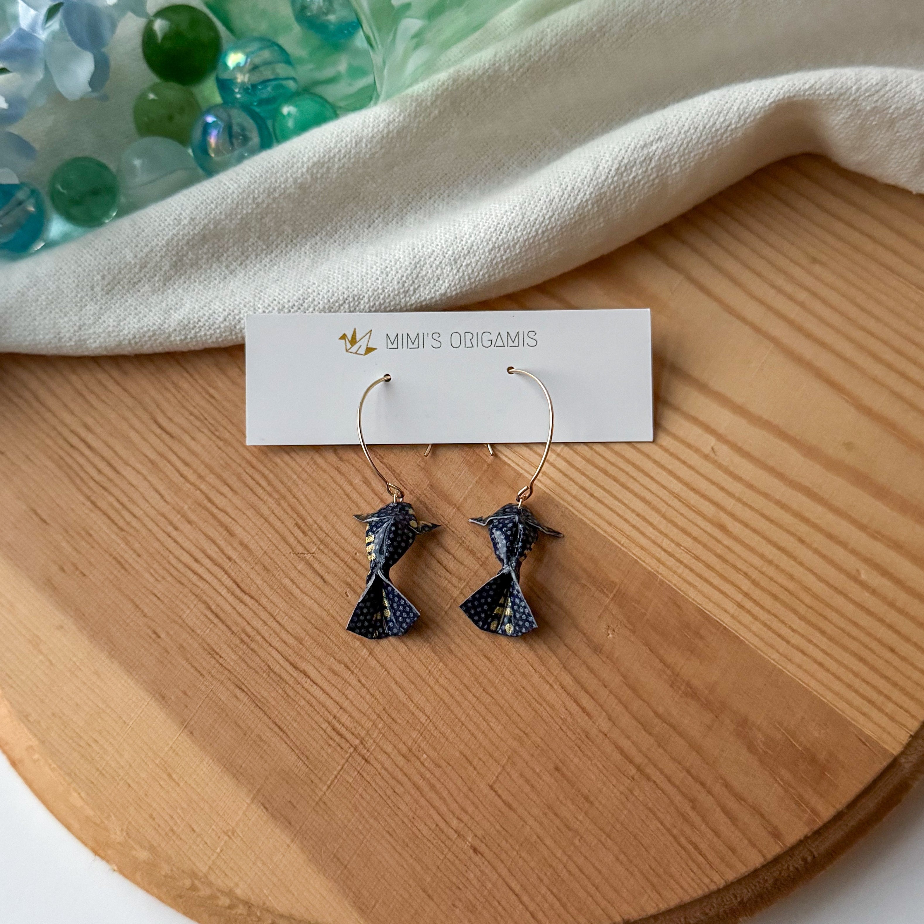 Pair of navy origami koi fish earrings on a wooden surface with a white branded card