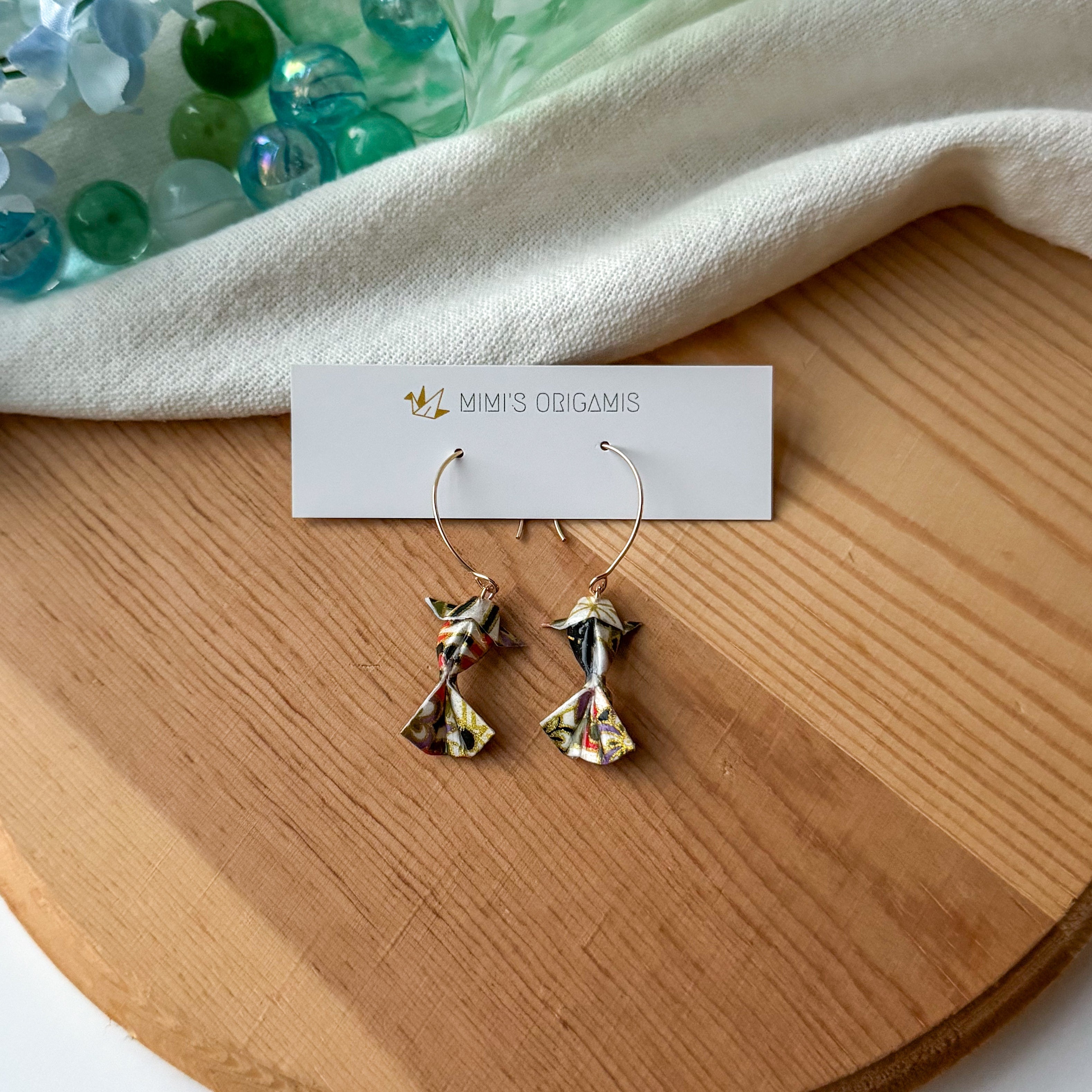 Pair of multicolored origami koi fish earrings on a wooden surface with a white branded card