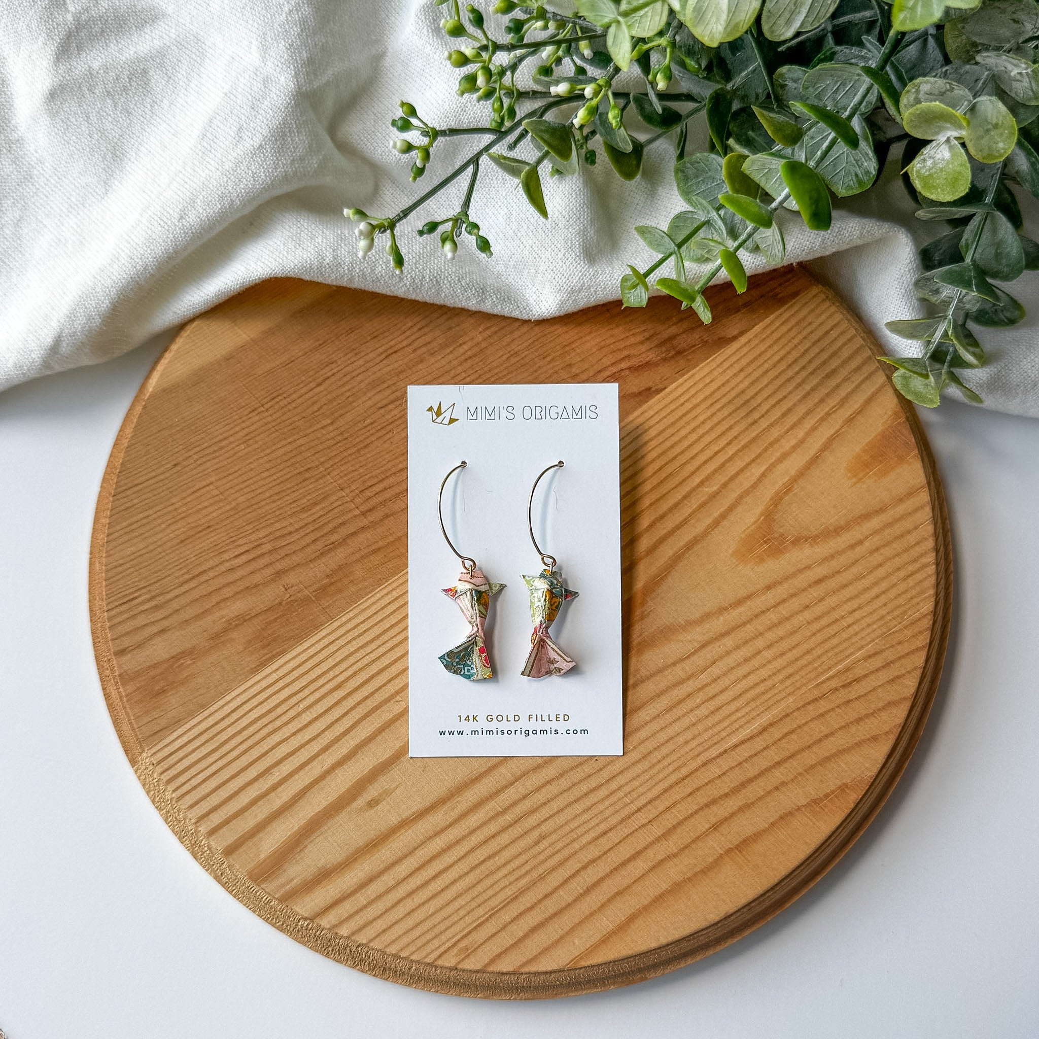 Pair of multicolored origami koi fish earrings on a wooden surface with a white branded card