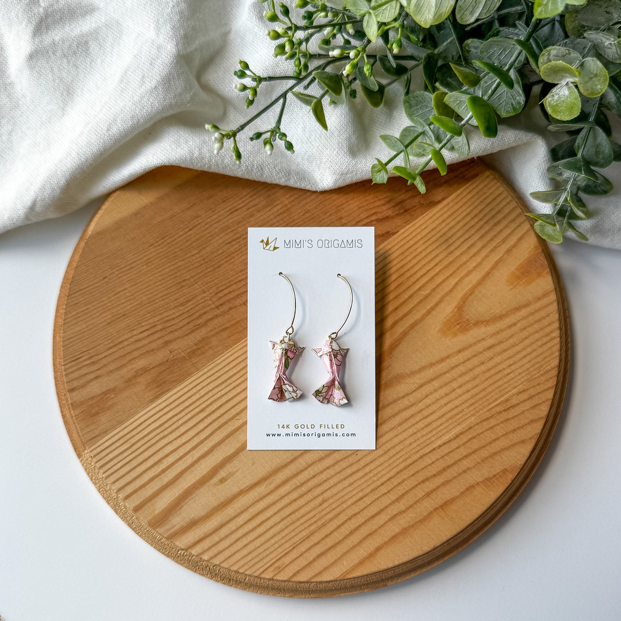 Pair of pink origami koi fish earrings on a wooden surface with a white branded card
