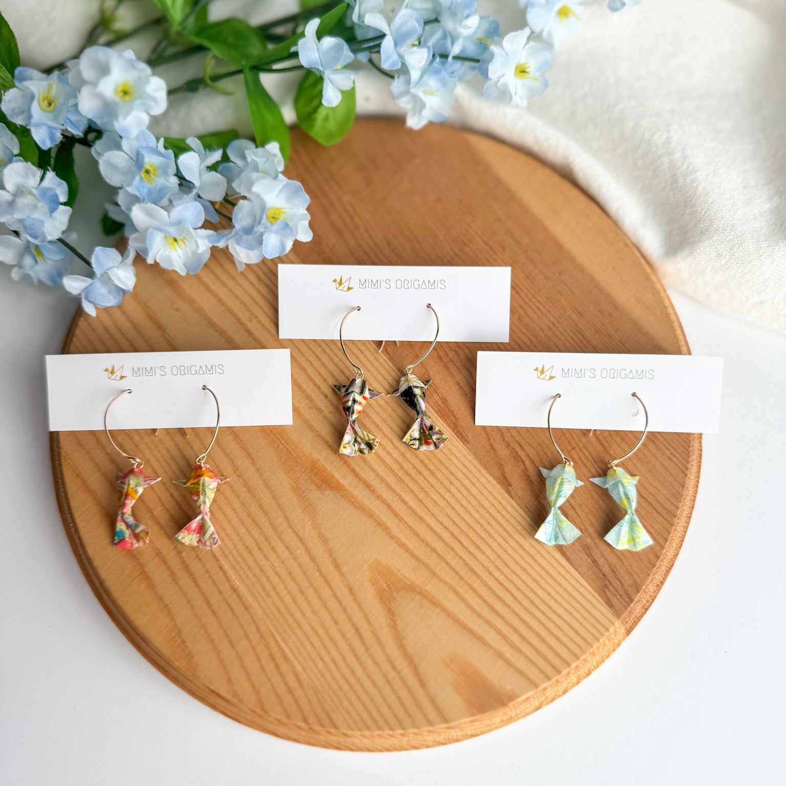 Set of origami koi fish earrings on wooden display with flowers and white fabric in the background