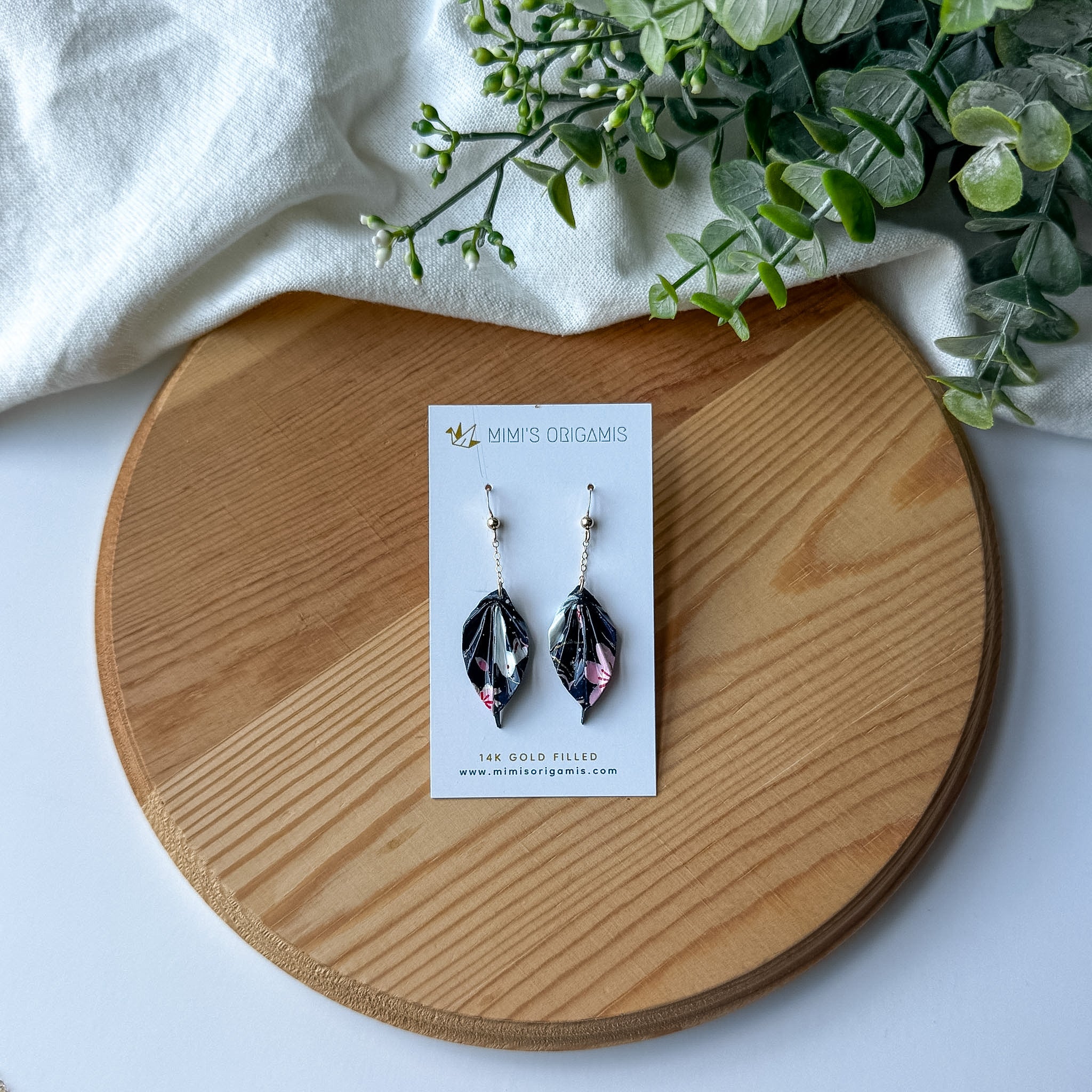 A pair of origami leaf earrings displayed on a white card, placed on a round wooden tray with a white cloth and green leaves in the background. 