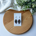 A pair of black origami leaf earrings displayed on a white card, placed on a round wooden tray with a white cloth and green leaves in the background. 