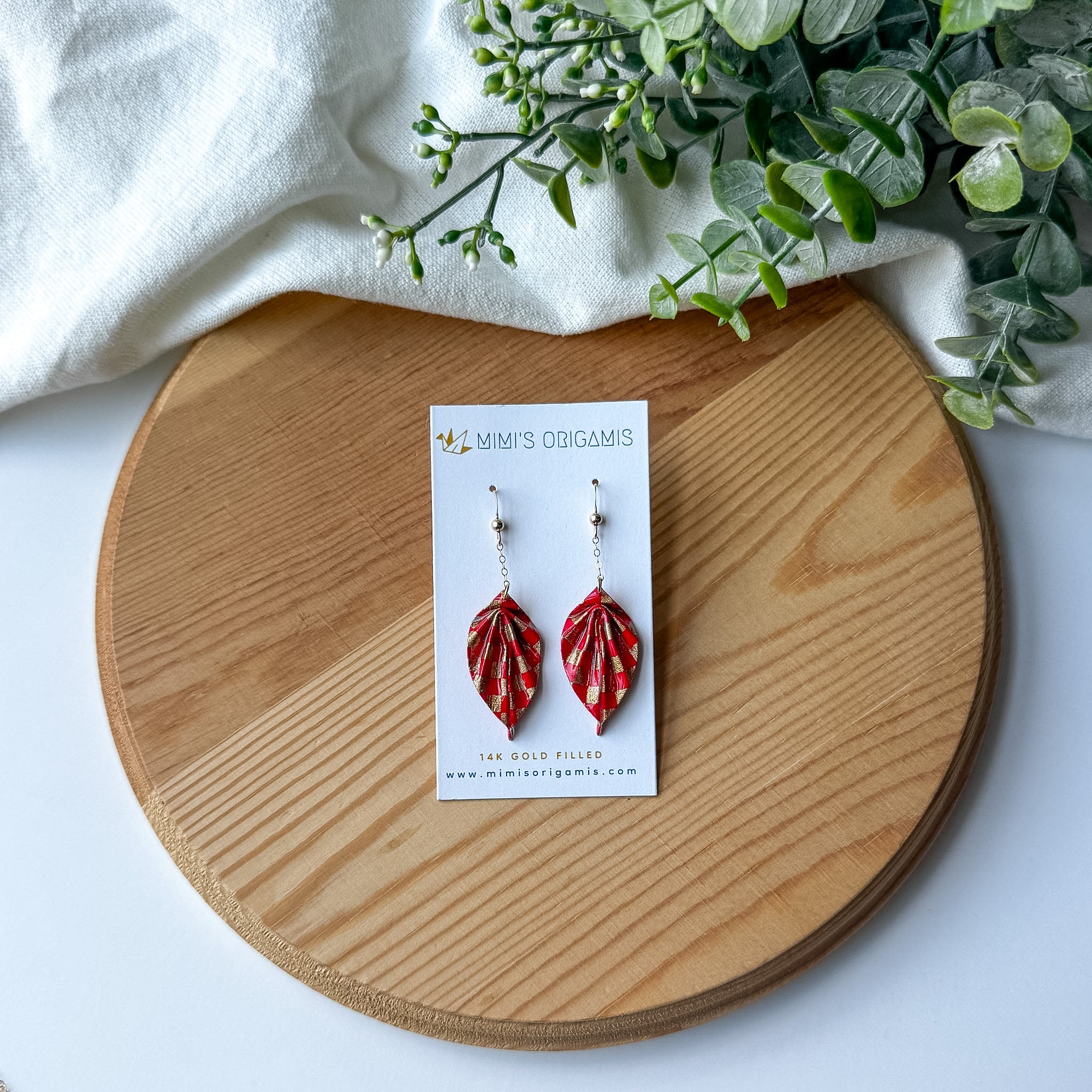 A pair of red and gold origami leaf earrings displayed on a white card, placed on a round wooden tray with a white cloth and green leaves in the background. 