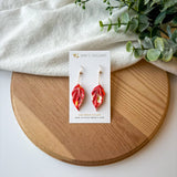 Colorful origami leaf earrings on a card with a wooden board and white fabric background