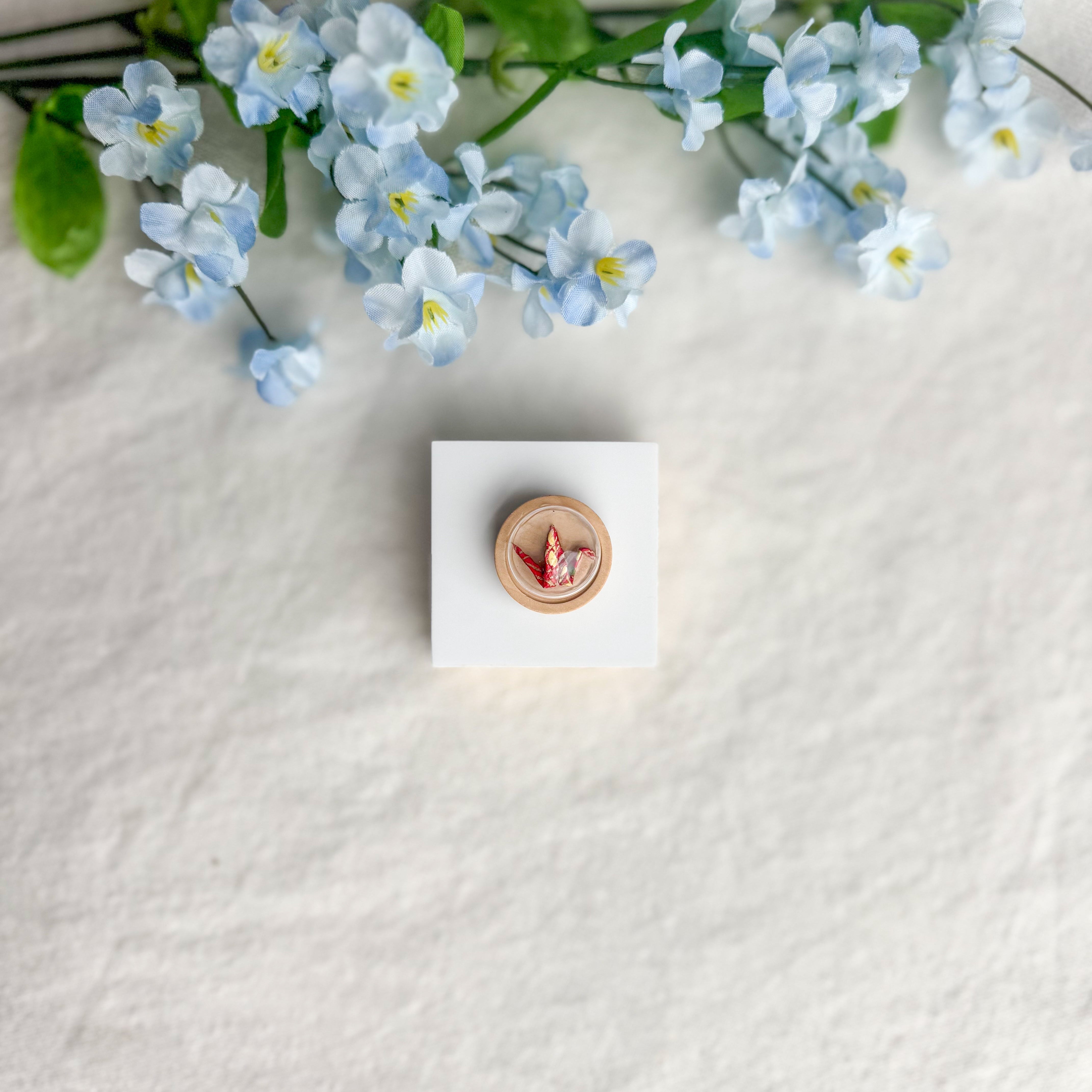 Round origami crane magnet on a white square surface next to blue flowers