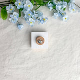 Round origami crane magnet on a white square surface next to blue flowers
