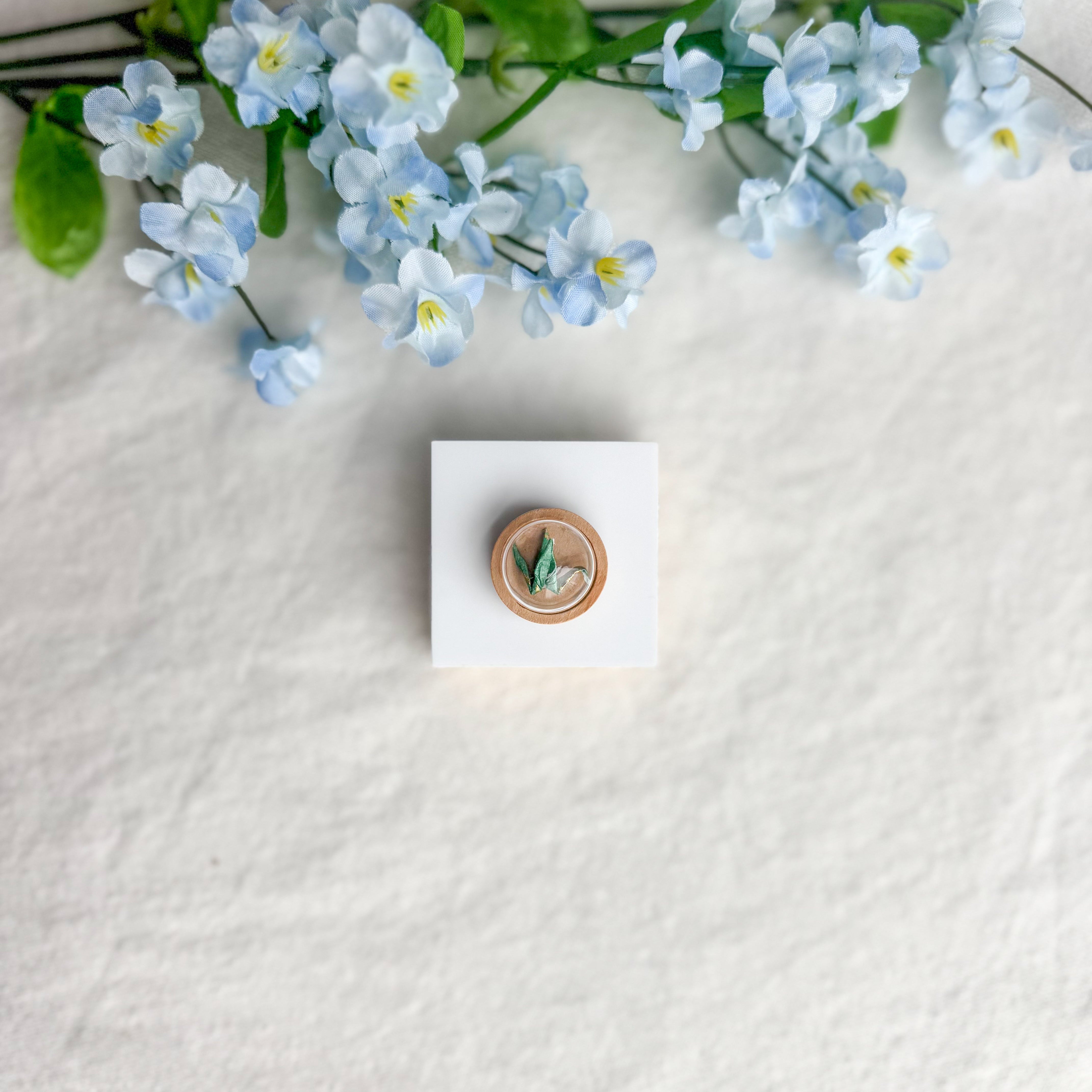 Round origami crane magnet on a white square surface next to blue flowers