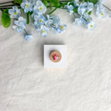 Round origami crane magnet on a white square surface next to blue flowers