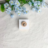 Round origami crane magnet on a white square surface next to blue flowers