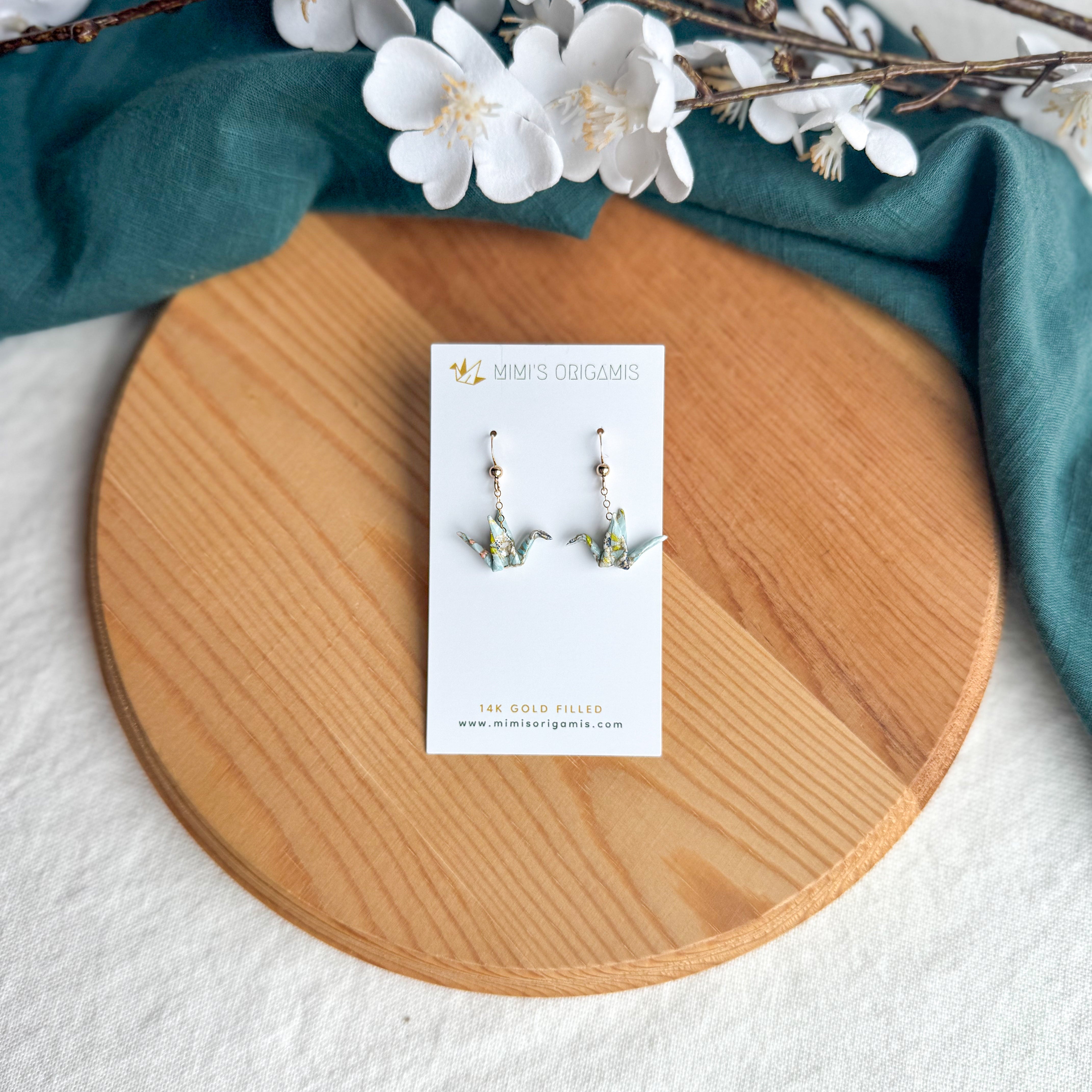 Origami crane earrings on a wooden surface with white flowers and green fabric