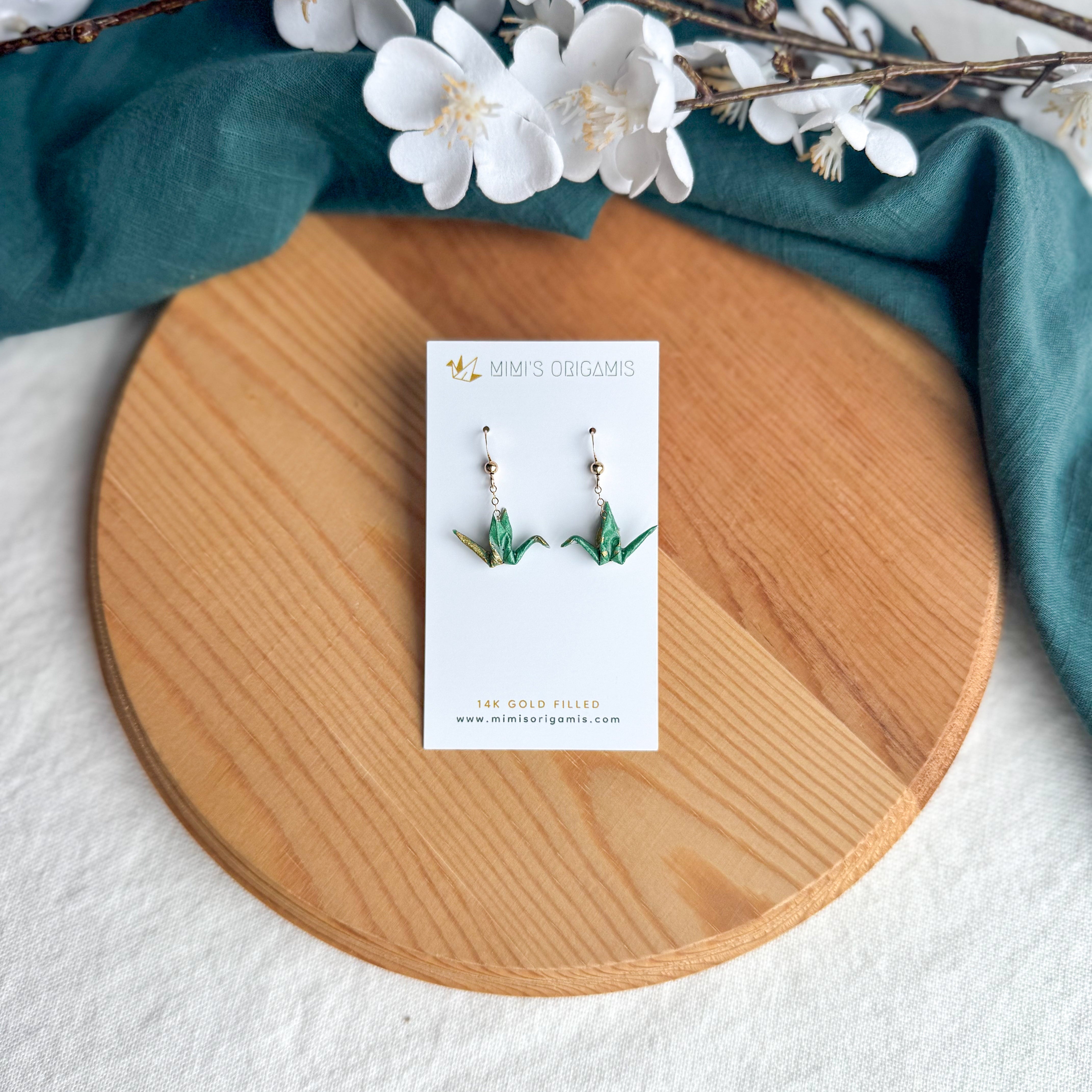 Origami crane earrings on a wooden surface with white flowers and green fabric
