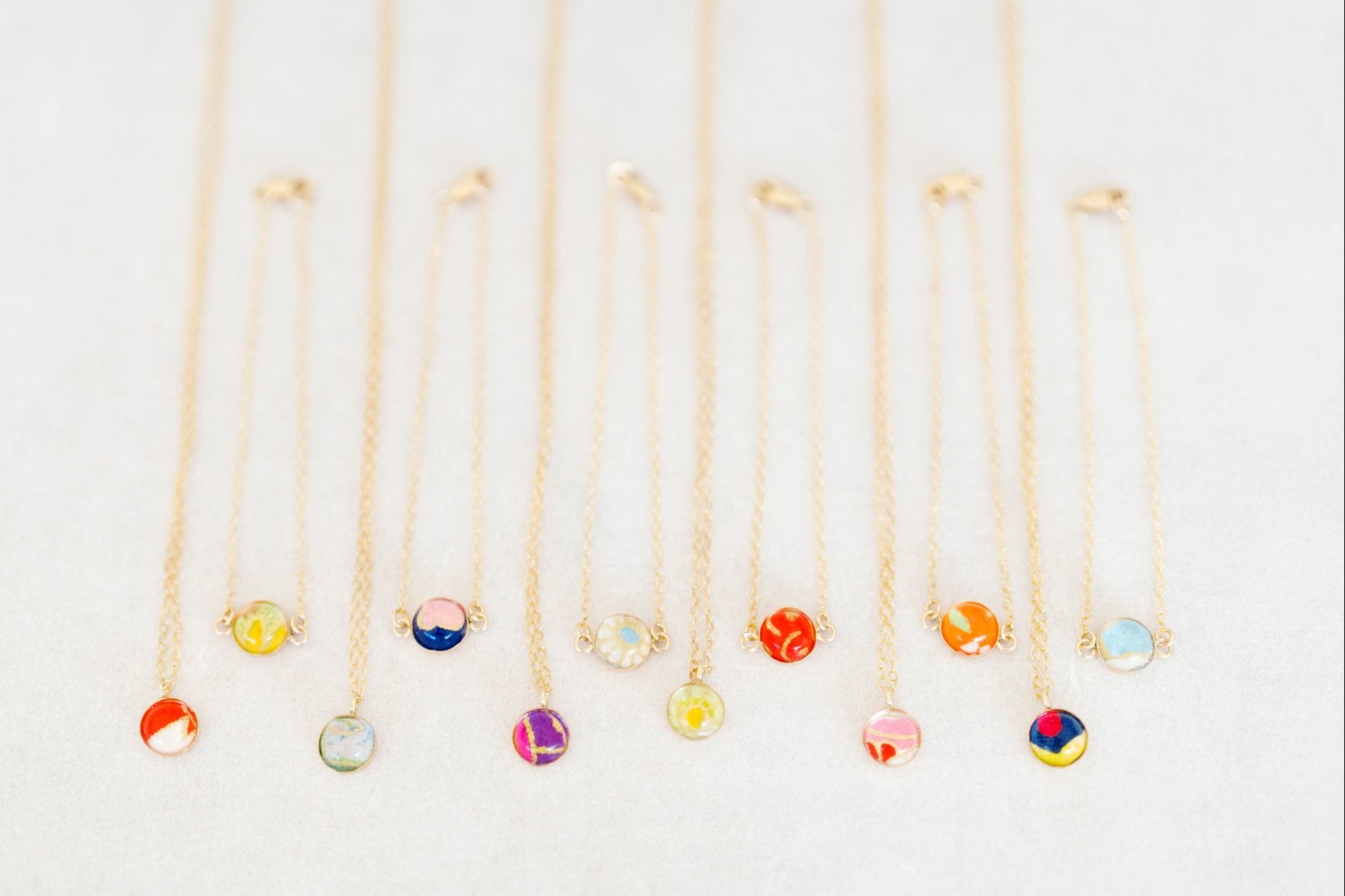 Six delicate gold chain necklaces and six bracelets with small round colorful gemstone pendants are arranged in a neat row on a light off-white textured background.