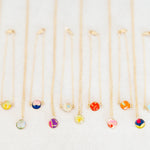 Six delicate gold chain necklaces and six bracelets with small round colorful gemstone pendants are arranged in a neat row on a light off-white textured background.