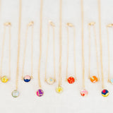 Six delicate gold chain necklaces and six bracelets with small round colorful gemstone pendants are arranged in a neat row on a light off-white textured background.