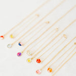 A set of delicate gold chain necklaces and bracelets with small, colorful flower-shaped pendants, arranged diagonally on a light off-white textured background.