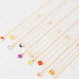 A set of delicate gold chain necklaces and bracelets with small, colorful flower-shaped pendants, arranged diagonally on a light off-white textured background.