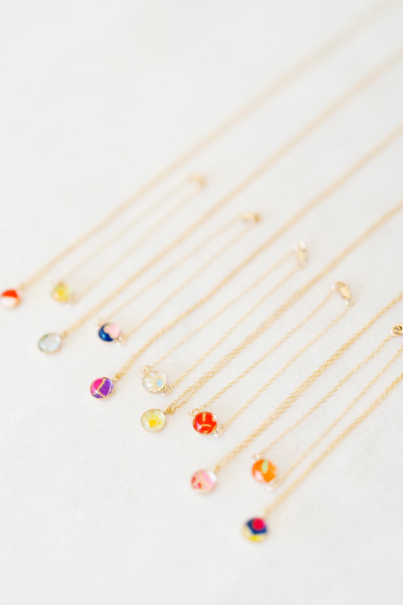 A set of delicate gold chain necklaces and bracelets with small, colorful flower-shaped pendants, arranged diagonally on a light off-white textured background.