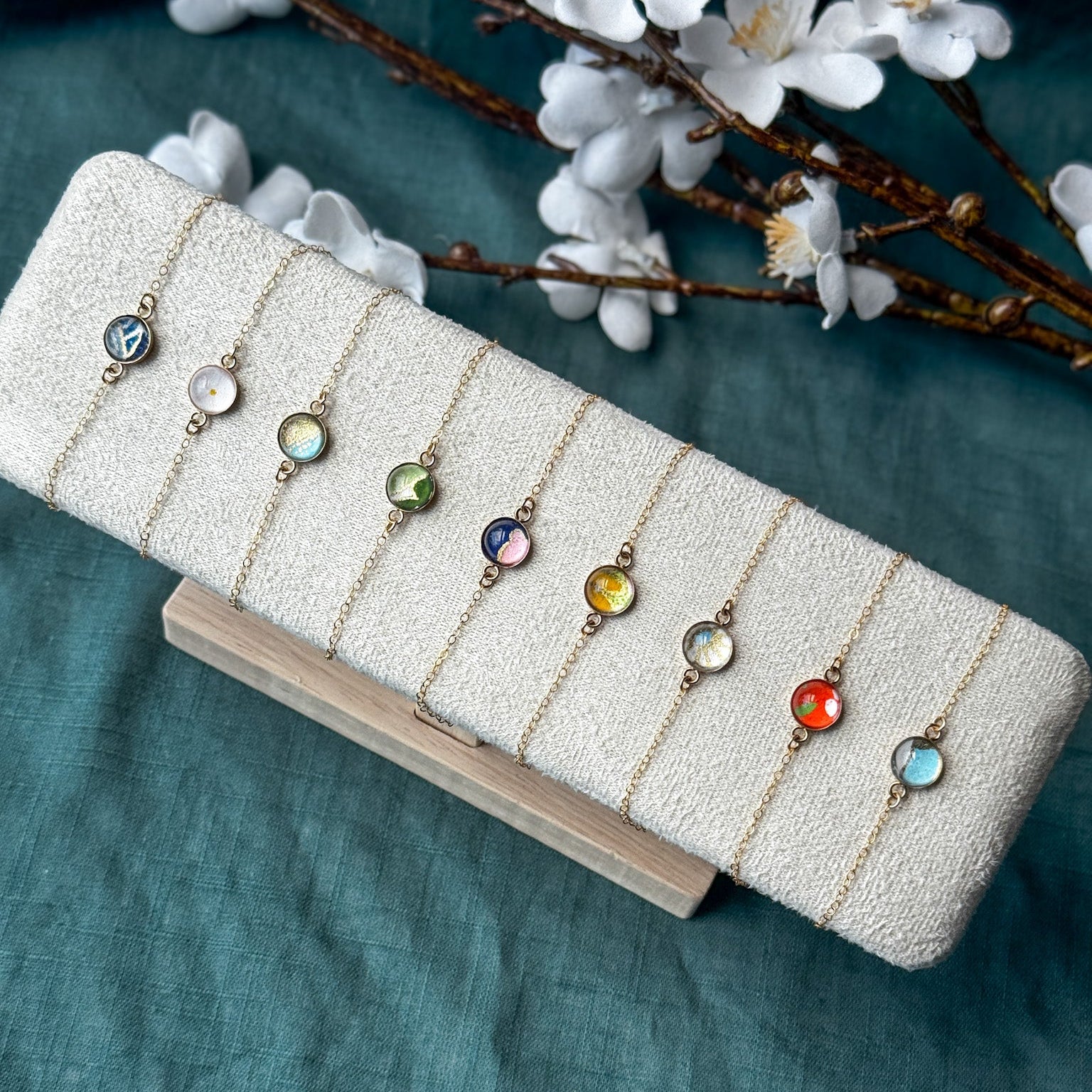 A set of nine delicate gold-toned necklaces with round, colorful gemstone pendants is displayed on a textured cream jewelry stand. The holder rests on a light wooden block over a teal fabric background, with white blossoms and brown branches framing the top of the image.