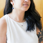 A woman with long dark hair wearing a white sleeveless top, wearing a colorful pendant necklace.