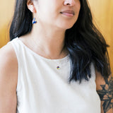 A woman with long dark hair wearing a white sleeveless top, wearing a colorful pendant necklace.