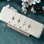 Four delicate gold-tone bracelets with round bezel-set domes are displayed on a textured cream jewelry stand. The holder rests on a light wooden block over a teal fabric background, with white blossoms and brown branches framing the top of the image.