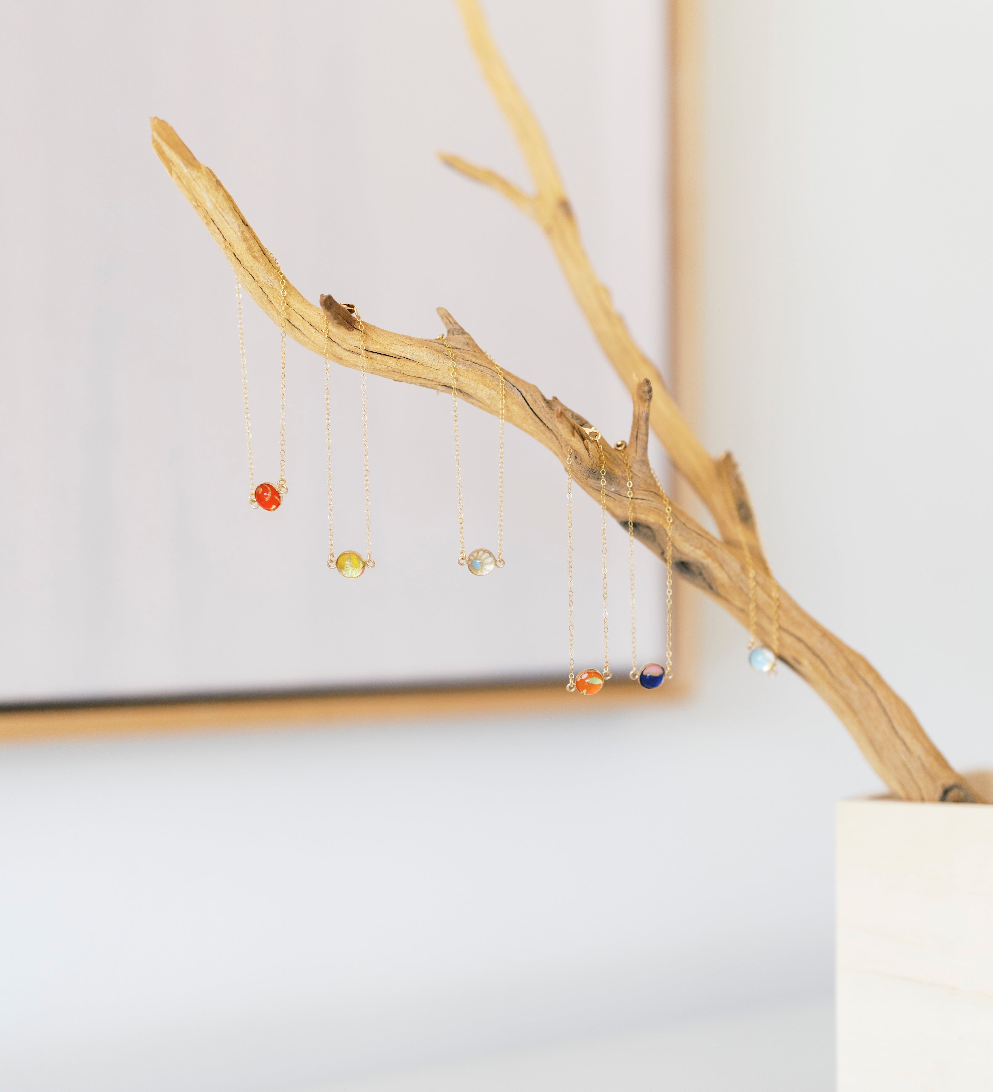 A set of delicate gold-tone bezel bracelets, hanging from a decorative driftwood branch stand in a white pot against a bright, softly blurred background.