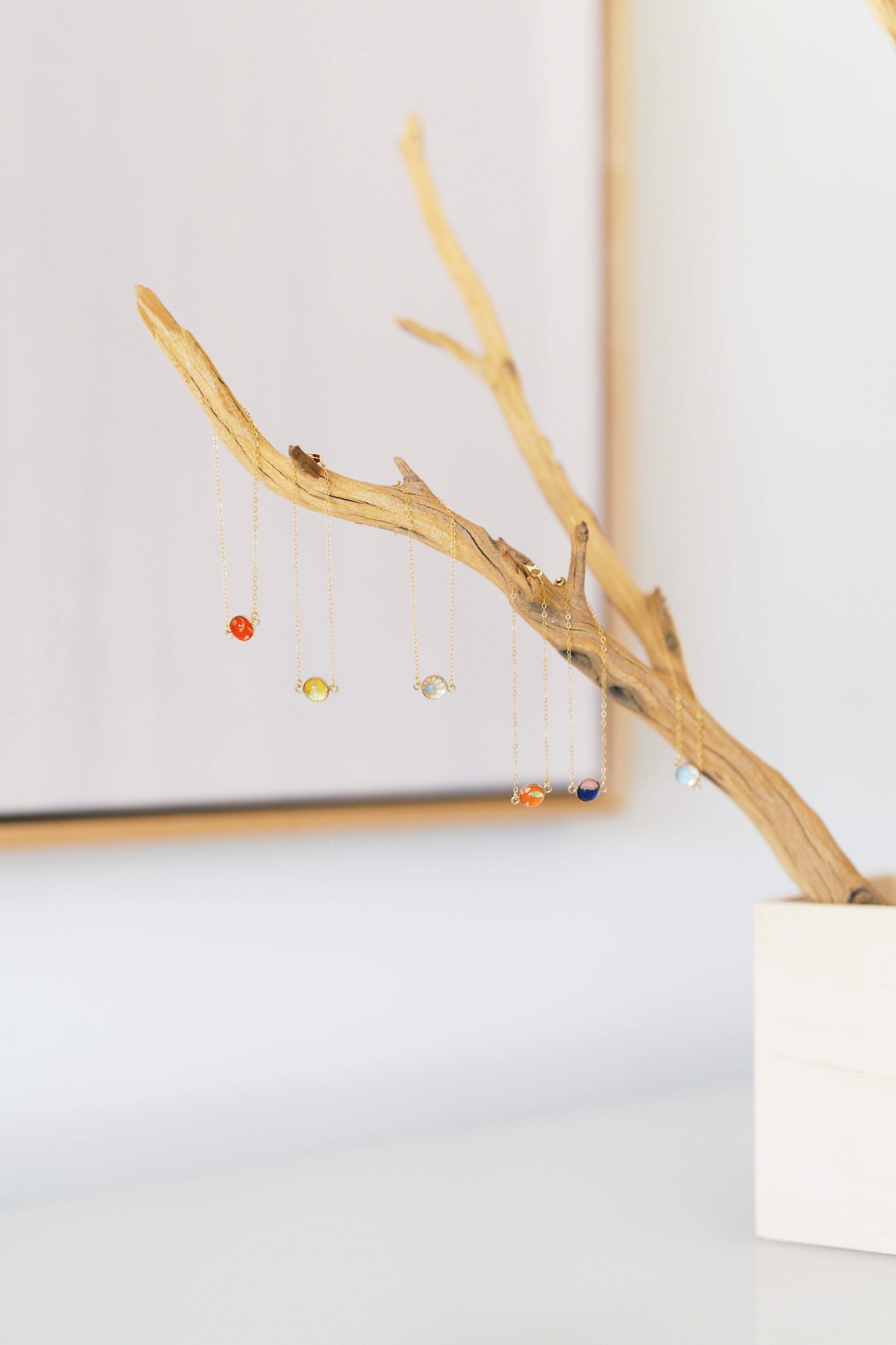 A set of delicate gold-tone bezel bracelets, hanging from a decorative driftwood branch stand in a white pot against a bright, softly blurred background.