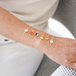 A close-up of a person's wrist wearing five delicate gold chain bracelets, each adorned with a small round domes in a different color. 
