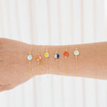 A close-up of a person's forearm wearing five delicate gold chain bracelets, each adorned with a small round stone in different colors, set against a soft white curtain background.