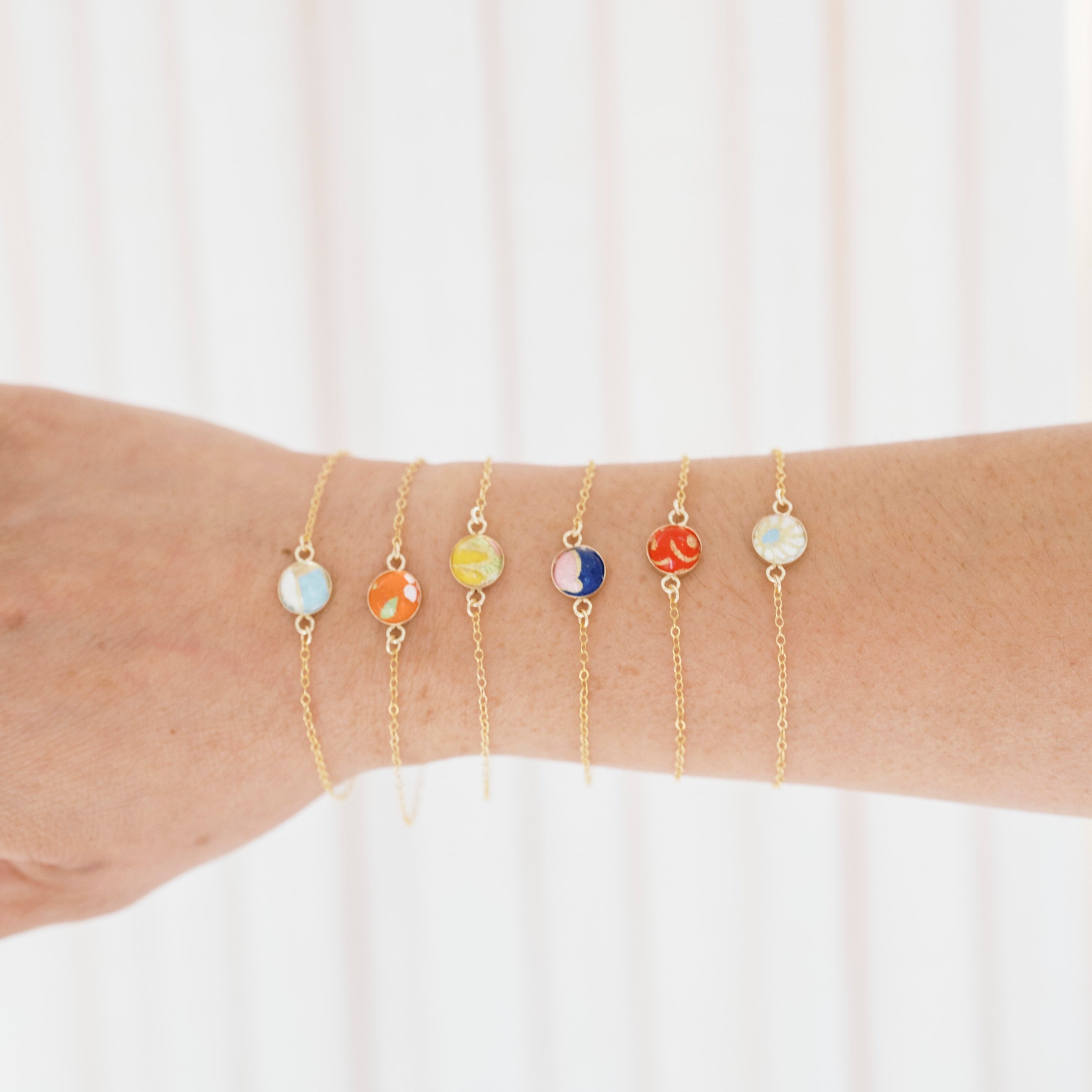 A close-up of a person's forearm wearing five delicate gold chain bracelets, each adorned with a small round stone in different colors, set against a soft white curtain background.
