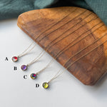 Four delicate gold-tone necklaces with small round gemstone pendants labeled A, B, C, and D, arranged on a curved wooden tray with a dark teal fabric backdrop.